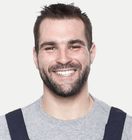 Smiling person with dark hair and beard, wearing a gray shirt and dark suspenders, on a white background